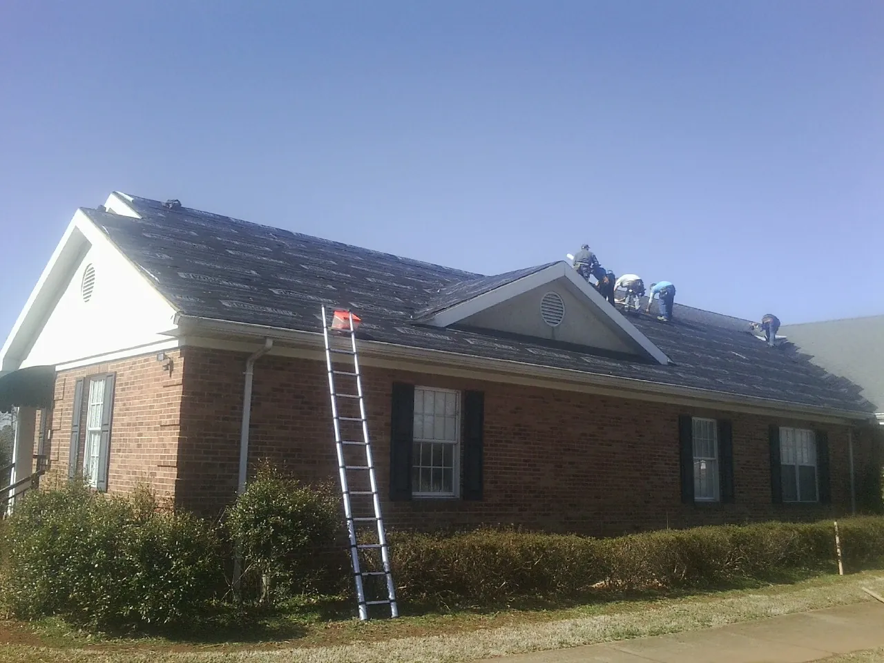 Crew tearing shingles off a brick ranch roof with a ladder set — Metal Roof Repair work by our crew in Highmount